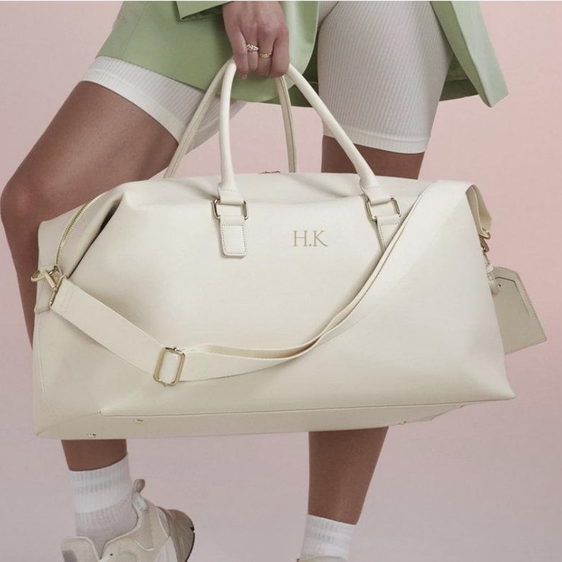 Puede incluir: Una bolsa de viaje de cuero blanco con una cremallera dorada y una correa dorada. La bolsa tiene las iniciales "H.K." bordadas en la parte delantera.