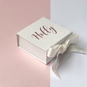 K&ouml;nnte beinhalten: Eine wei&szlig;e Geschenkbox mit einem wei&szlig;en Satinband, das zu einer Schleife gebunden ist. Auf der Oberseite der Box ist der Name "Holly" in ros&eacute;goldener Schrift gedruckt.