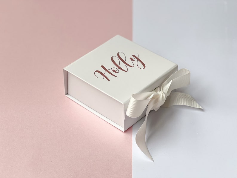K&ouml;nnte beinhalten: Eine wei&szlig;e Geschenkbox mit einem wei&szlig;en Satinband, das zu einer Schleife gebunden ist. Auf der Oberseite der Box ist der Name "Holly" in ros&eacute;goldener Schrift gedruckt.