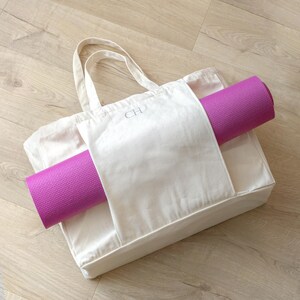 May include: A white canvas tote bag with a monogram "CH" embroidered on the front. The bag has a separate pocket on the side for a rolled up purple yoga mat.