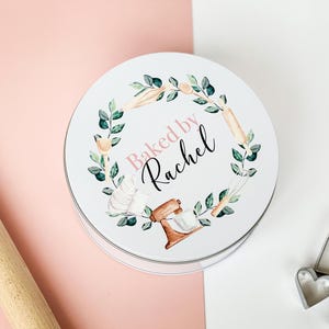 May include: White round tin with a green wreath design featuring baking tools and the text "Baked by Rachel".