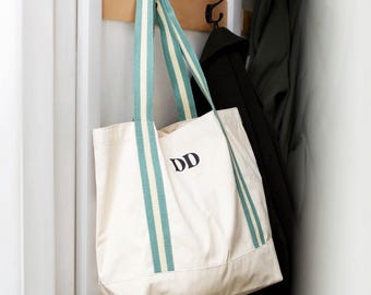 May include: A white canvas tote bag with green and white striped straps. The bag has the initials "DD" embroidered on the front.