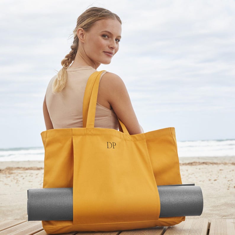 May include: A yellow canvas tote bag with a black monogram "DP" on the front. The bag has a pocket on the side to hold a grey yoga mat.