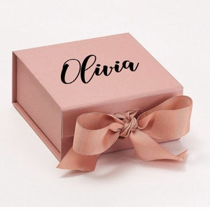 K&ouml;nnte beinhalten: Eine rosa Geschenkbox mit einem Satinband, das zu einer Schleife gebunden ist. Auf der Oberseite der Box ist der Name "Olivia" in schwarzer Kursivschrift aufgedruckt.