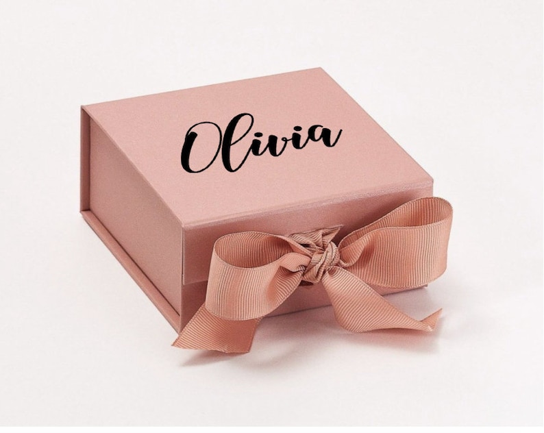 K&ouml;nnte beinhalten: Eine rosa Geschenkbox mit einem Satinband, das zu einer Schleife gebunden ist. Auf der Oberseite der Box ist der Name "Olivia" in schwarzer Kursivschrift aufgedruckt.
