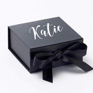 K&ouml;nnte beinhalten: Eine schwarze Geschenkbox mit einem wei&szlig;en Band und dem Namen "Katie" auf der Oberseite.
