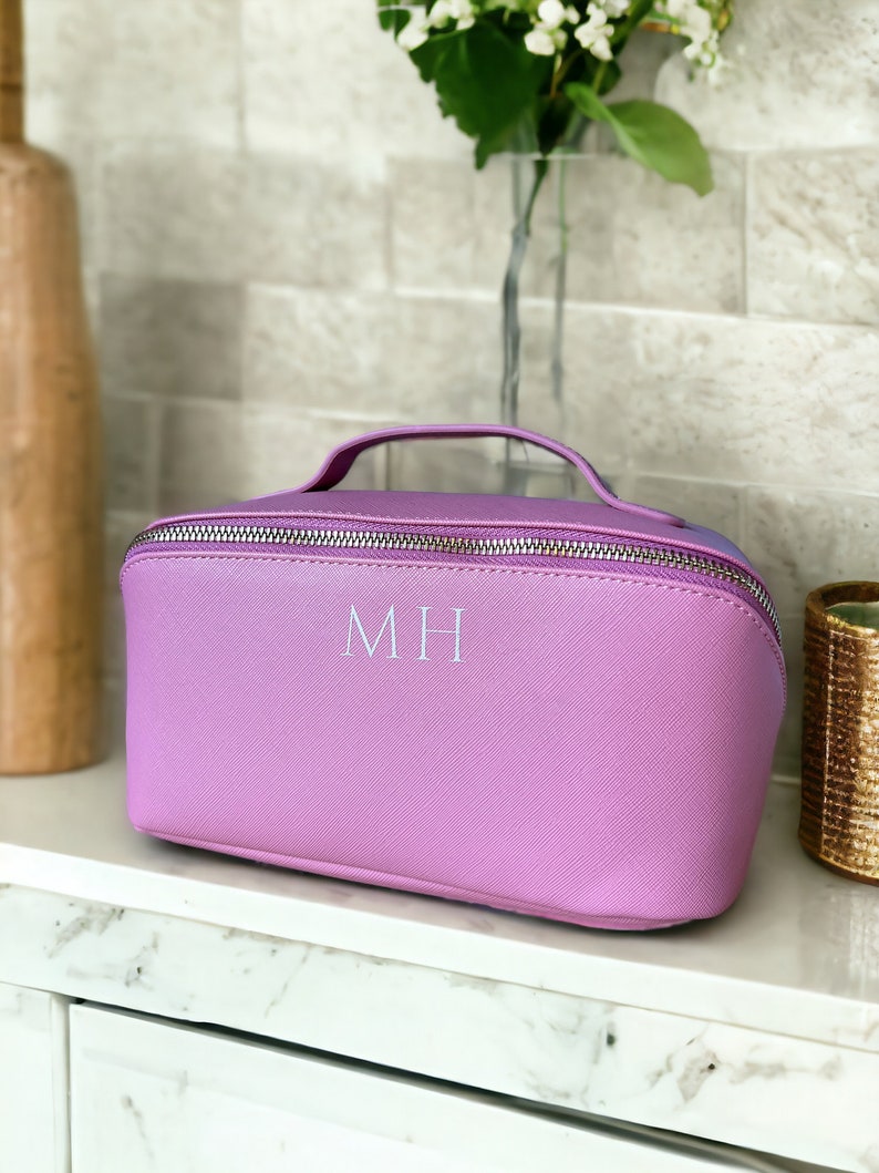 May include: A lavender-colored cosmetic bag with a zipper closure and a silver handle. The bag has the initials "MH" embroidered on the front in white.