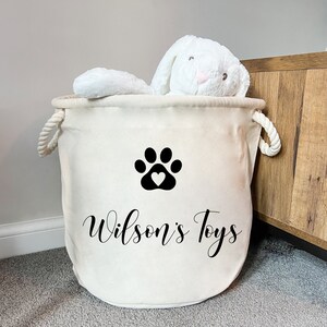May include: A cream-colored fabric storage basket with two white rope handles. The front features a black paw print graphic with a heart in the center and the text "Wilson's Toys" in a black script font. A white plush rabbit toy sits inside.