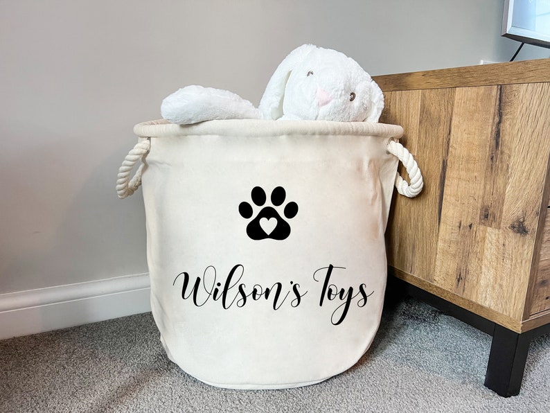 May include: A cream-colored fabric storage basket with two white rope handles. The front features a black paw print graphic with a heart in the center and the text "Wilson's Toys" in a black script font. A white plush rabbit toy sits inside.