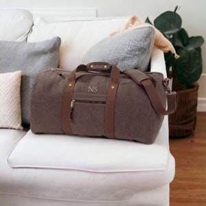 Puede incluir: Una bolsa de lona marr&oacute;n con detalles de cuero y un bolsillo con cremallera. La bolsa tiene una cremallera de metal dorado y una hebilla de metal dorado. La bolsa est&aacute; monogramada con las iniciales "NS".