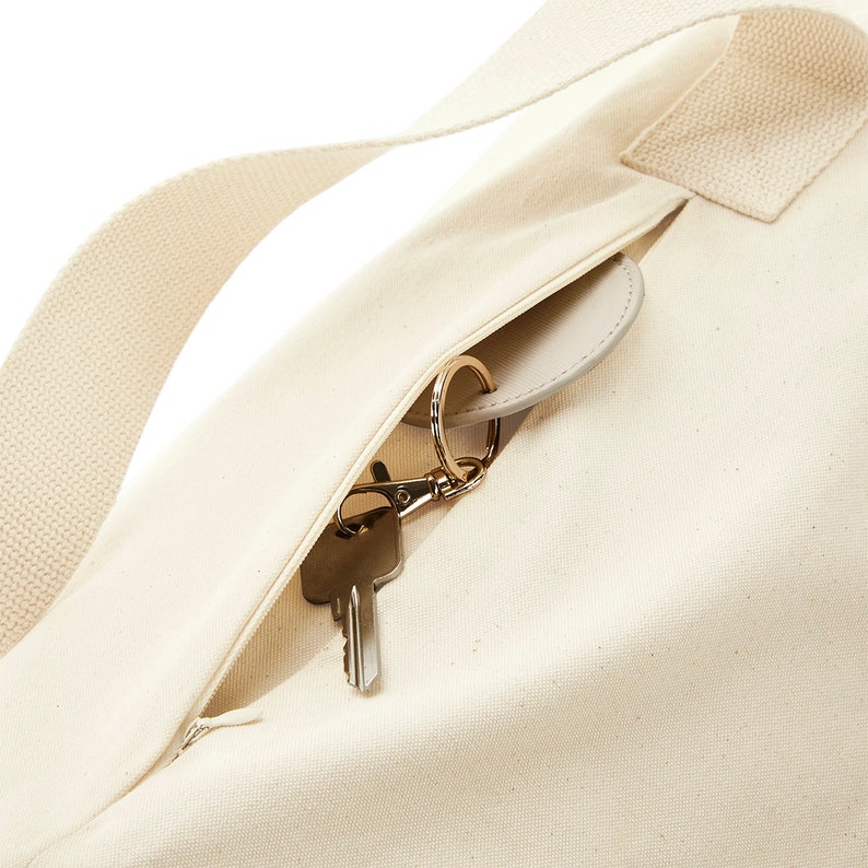 May include: A close-up of a cream-colored canvas tote bag with a zippered pocket. Keys are visible inside the pocket.