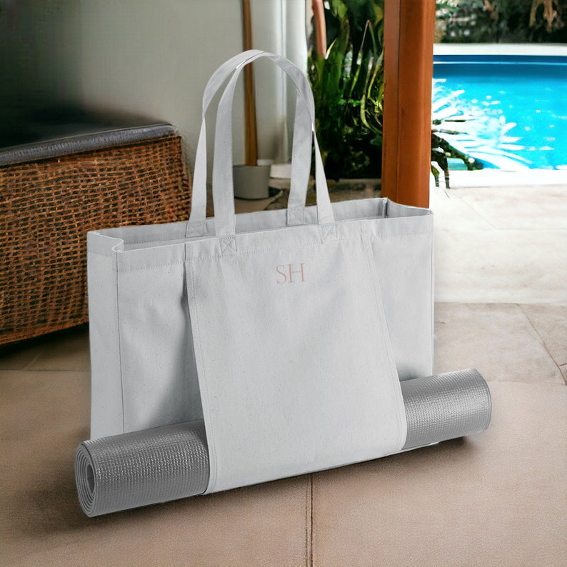 May include: A white canvas tote bag with a gray yoga mat tucked into the side pocket. The bag has the initials "SH" embroidered on the front.