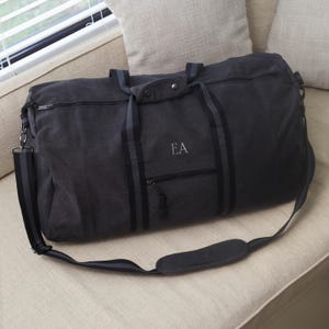 Puede incluir: Una bolsa de lona negra con rayas negras y grises y un bolsillo con cremallera. La bolsa tiene una correa para el hombro y las iniciales "EA" bordadas en la parte delantera.