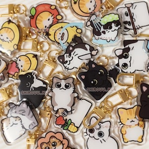 May include: A collection of clear acrylic keychains featuring various illustrated cats in different poses and colors, including orange tabby, black, white, and gray patterns. Each keychain is attached to a gold-toned metal lobster clasp.