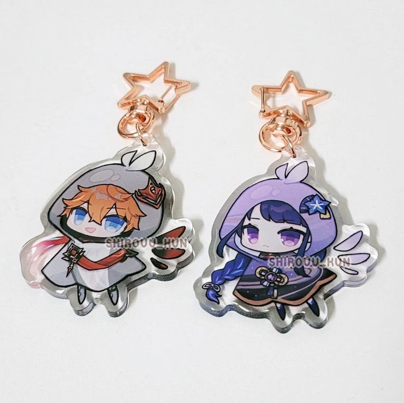 May include: Two acrylic charms featuring chibi-style anime characters with star-shaped rose gold clasps. The left charm depicts a character with orange hair in a grey hood, while the right charm shows a character with purple hair in a purple hood.