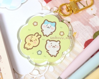 Pode incluir: Um porta-chaves de acrílico em forma de flor com um fundo verde claro e três personagens de desenhos animados: uma criatura branca com orelhas azuis, um coelho amarelo e um urso branco. O texto SHIROUU_KUN está impresso com um fecho metálico dourado.