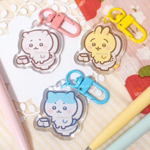 Peut inclure: Trois porte-clés en acrylique transparent représentant des personnages de dessin animé assis sur des toilettes avec du papier toilette. Un personnage est blanc avec un fermoir rose, un jaune avec un fermoir jaune, et un bleu avec un fermoir bleu. Texte SHIROUU_KUN.