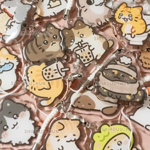 May include: A collection of clear acrylic charms featuring various illustrated cats in different poses and outfits, including cats with bubble tea, hats, and scarves. Each charm has a white cord loop and the text SHIROUU_KUN printed on the surface.