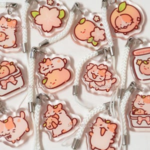 May include: A collection of clear acrylic charms featuring illustrations of cats and peaches with cherry blossoms. Each charm is attached to a white braided cord with a silver metal clasp. The text SHIROUU_KUN is printed on the charms.