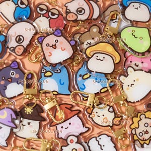 May include: A collection of clear acrylic keychains featuring various cartoon characters, including animals, mushrooms, and ghosts, each with a gold-toned metal lobster clasp. The text SHIROUU_KUN is printed in small letters across several of the charms.
