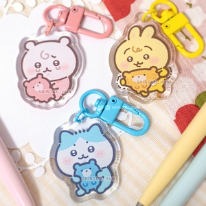 May include: Three acrylic keychains featuring cartoon characters holding smaller figures. One is pink with a pink clasp, one is yellow with a yellow clasp, and one is blue with a blue clasp. Each keychain has the text SHIROUU_KUN printed on the surface.