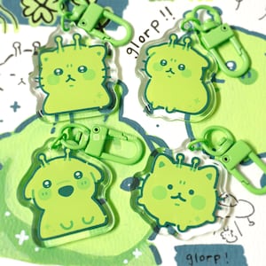 以下が含まれることがあります： 緑色の宇宙人のようなキャラクターのキーホルダー4つと緑色の留め金。キーホルダーは、大きな目と頬の赤みが特徴的な漫画風のデザインです。背景には、水彩画のような緑と青の形と「glorp !!」の文字があります。
