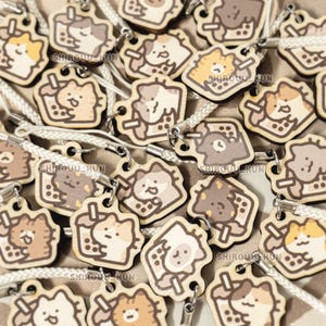 May include: A collection of wooden charms featuring a cartoon cat holding a bubble tea drink with the text SHIROUU_HUN printed on each piece. The charms are light brown and cream colored with a white braided cord attached to a metal jump ring.