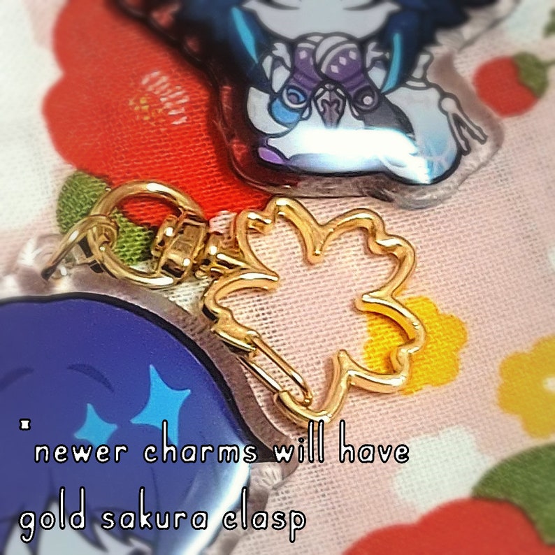 Puede incluir: Dos dijes de acr&iacute;lico con personajes de estilo anime y un cierre de metal dorado con forma de flor de cerezo. El texto en la imagen dice: newer charms will have gold sakura clasp. Los dijes son transparentes con dise&ntilde;os coloridos.