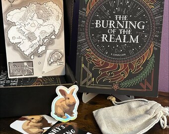 May include: A book titled The Burning of the Realm by S. Lynn Cox with a black cover and gold flame graphics, a map of the realm, a small beige drawstring pouch, a rabbit sticker, and a rectangular bookmark featuring rabbits and the text Henry's Resting.