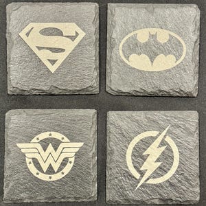 May include: Four square slate coasters featuring superhero logos. The set includes the Superman logo, the Batman logo, the Wonder Woman logo, and the Flash logo. The logos are in a light color against the dark gray slate.