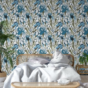 May include: A white bed with a light blue floral patterned wallpaper behind it. The bed has a white duvet and a few pillows. There is a wooden bench in front of the bed and a small table with a plant on it to the right.