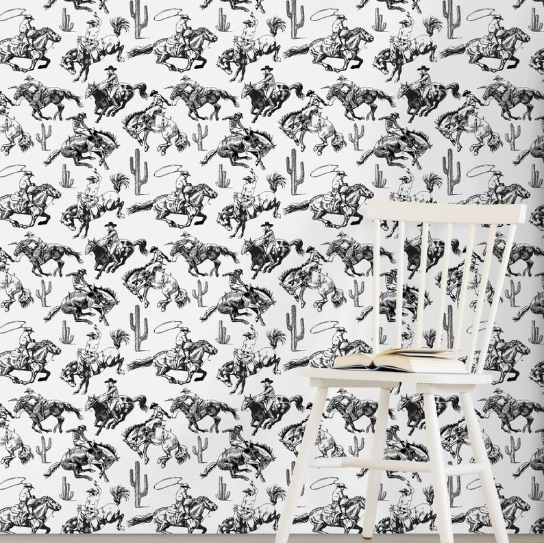 May include: Black and white repeating pattern wallpaper featuring cowboys riding horses with lassoes. The pattern includes cacti and rope circles. A white chair with a book on it is in the foreground.