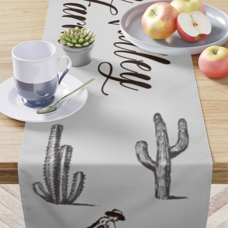 May include: A white table runner with a black and white cactus design and the text "Farm Valley" in black cursive writing. The table runner is on a wooden table with a cup, saucer, and a small potted plant.