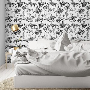 May include: Black and white wallpaper featuring a repeating pattern of cowboys riding horses. The wallpaper is displayed on a white wall behind a white bed with white bedding.