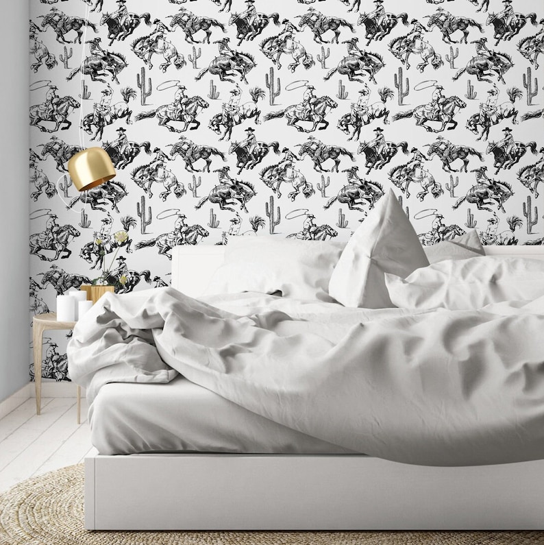 May include: Black and white wallpaper featuring a repeating pattern of cowboys riding horses. The wallpaper is displayed on a white wall behind a white bed with white bedding.