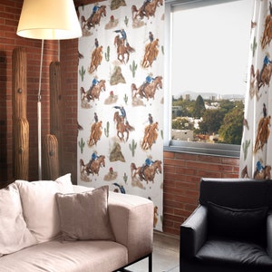 May include: White curtains with a repeating pattern of cowboys on horseback, cacti, and desert plants. The curtains are hanging in front of a window, and the room has a modern, rustic style with a brick wall and a beige sofa.
