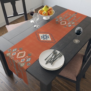 May include: A rust-colored table runner with a geometric pattern in shades of blue, gray, and white. The runner is laid on a dark wood table with a bowl of apples, two wine glasses, and a stack of white plates.