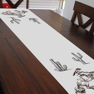 May include: White table runner with a brown cactus and cowboy riding a horse design.