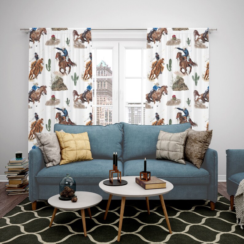 May include: White window curtains with a repeating watercolor pattern of cowboys riding horses, cacti, and desert landscapes. The curtains are hanging in front of a window, with a blue sofa and coffee tables in front of them.