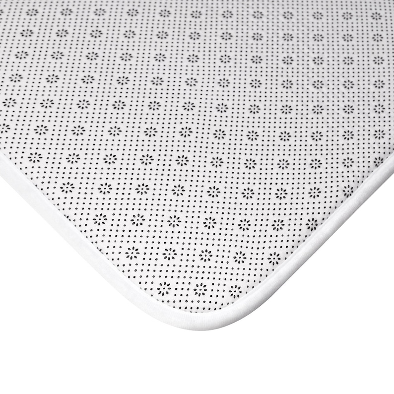 Puede incluir: Primer plano de la parte inferior blanca de una alfombra de ba&ntilde;o. La alfombra tiene un borde blanco y una superficie antideslizante negra texturizada.