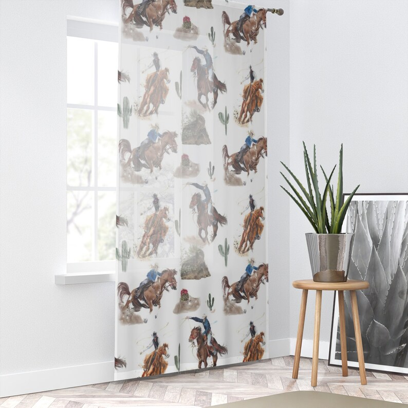 May include: Sheer white curtain panel featuring a repeating watercolor pattern of cowboys riding horses, cacti, and desert landscapes. The design includes cowboys in blue shirts and brown horses in action poses. The curtain is hanging in front of a window.