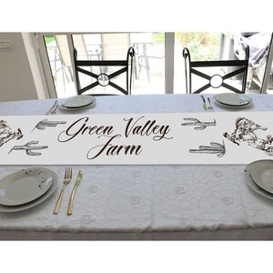 May include: White table runner with a brown 'Green Valley Farm' text and a black and white illustration of a cactus and a horse. The runner is on a white table with two chairs.