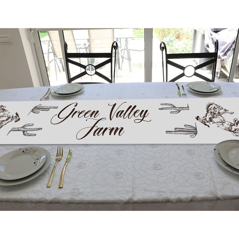 May include: White table runner with a brown 'Green Valley Farm' text and a black and white illustration of a cactus and a horse. The runner is on a white table with two chairs.