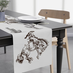 May include: White table runner with a brown cowboy riding a bucking horse and cactus illustrations. The text "Howdy" is written in cursive at the top of the runner.