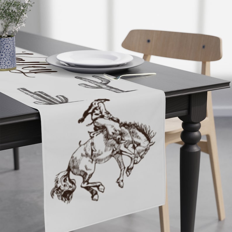 May include: White table runner with a brown cowboy riding a bucking horse and cactus illustrations. The text "Howdy" is written in cursive at the top of the runner.
