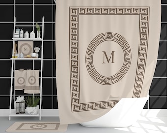 May include: A beige shower curtain with a brown Greek key border and a central circle with the letter "M". Matching towels, a bath mat, and a framed print are also visible, creating a coordinated bathroom set. The bathroom has black tile.
