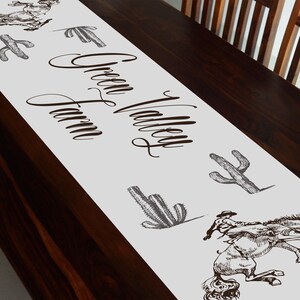 May include: White table runner with the text "Green Valley Farm" in brown cursive script. The runner is decorated with black and white illustrations of cacti and a cowboy riding a horse.