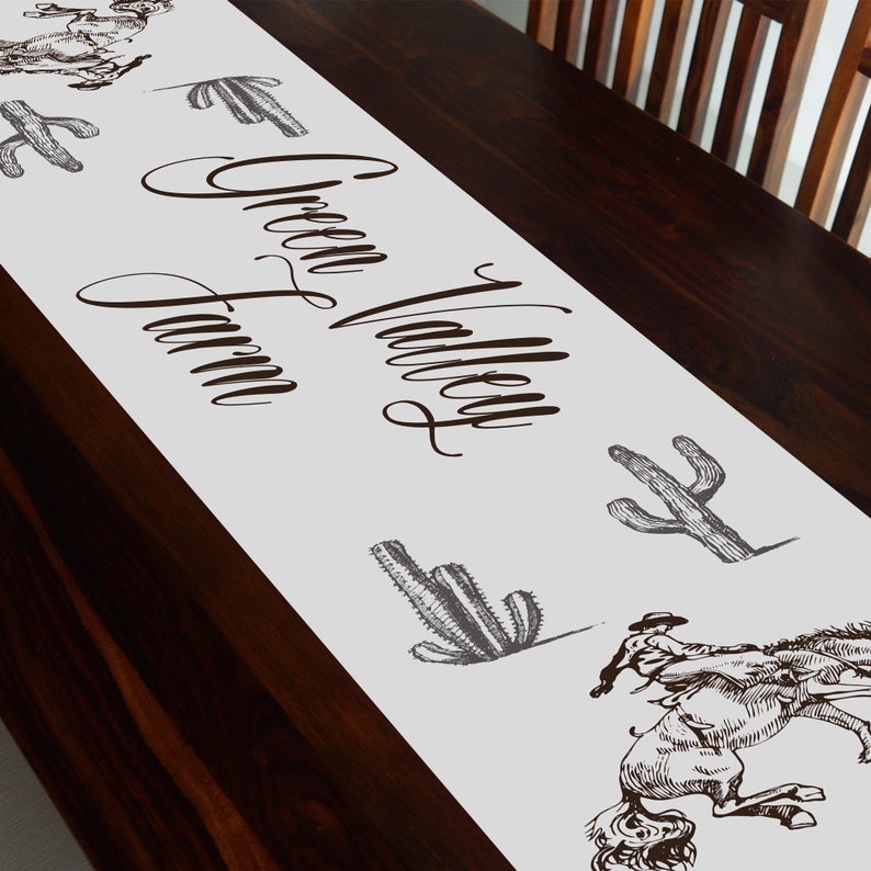 May include: White table runner with the text "Green Valley Farm" in brown cursive script. The runner is decorated with black and white illustrations of cacti and a cowboy riding a horse.