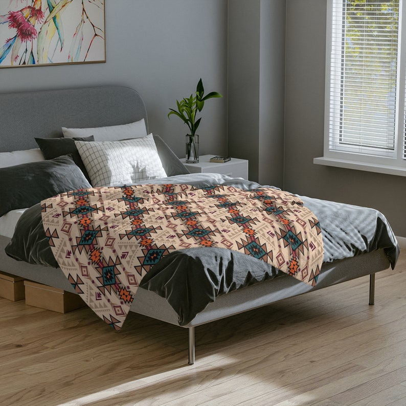 K&ouml;nnte beinhalten: Eine beigefarbene Tagesdecke mit einem geometrischen Muster in Orange-, Rot-, Blau- und Braunt&ouml;nen. Die Decke liegt auf einem Bett mit grauer Bettw&auml;sche.