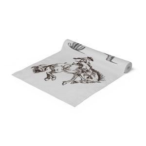 May include: A white table runner with a brown illustration of a cowboy riding a bucking horse. The runner also features two black and white illustrations of cacti.